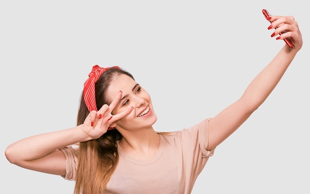 Giovane donna europea in abiti casual e fascia rossa che si autoritratto su un muro bianco dello studio Femmina felice che sorride e si fa un selfie sul suo smartphone