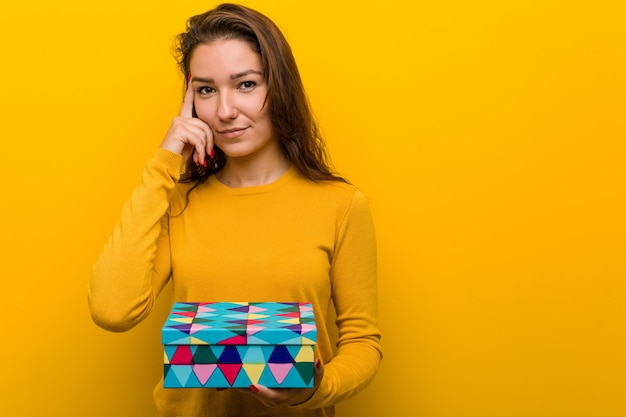 Giovane donna europea in possesso di un regalo che punta il suo tempio con il dito, pensando, incentrato su un compito.