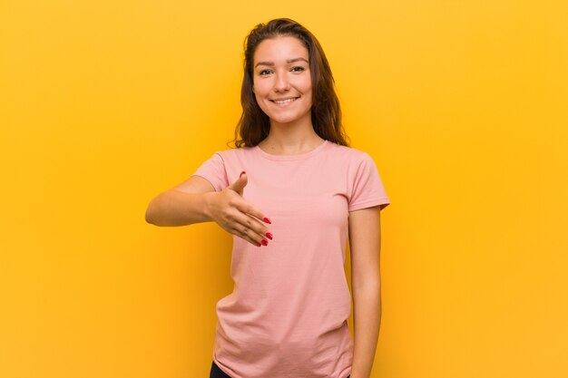 Giovane donna europea isolata sopra la parete gialla che allunga mano alla macchina fotografica nel gesto di saluto