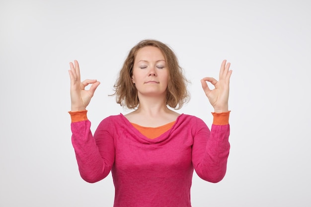 Giovane donna europea sorridente in maglione rosa che medita tenendo le mani in un gesto di yoga sentendosi calma e positiva