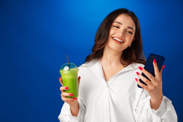 Giovane donna felice attraente che tiene una tazza di frullato verde e usa il suo smartphone contro b...