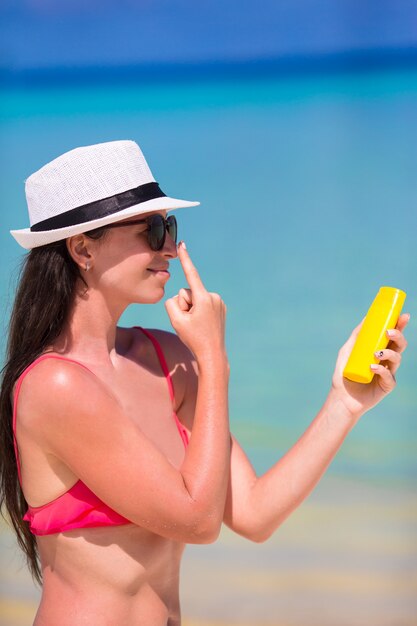 Giovane donna felice che applica lozione solare sul suo naso sulla spiaggia bianca