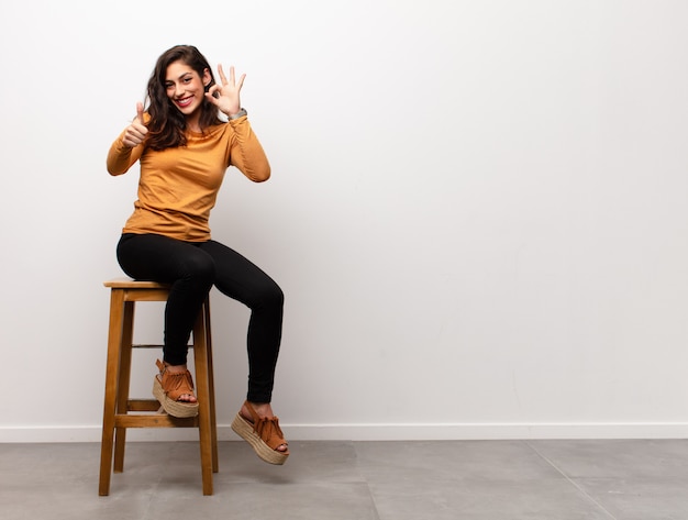 Giovane donna felice che fa gesto giusto, sedendosi su una sedia accanto a copyspace