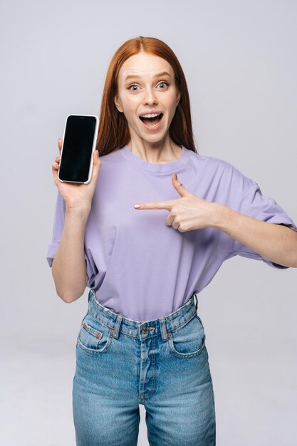 Giovane donna felice che indossa abiti casual eleganti che puntano con il dito sullo schermo mobile vuoto. Modello da donna graziosa con capelli rossi che mostra emotivamente le espressioni facciali in studio, copyspace.