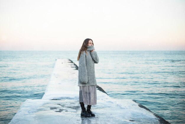 Giovane donna felice che indossa un maglione di lana in piedi sulla spiaggia in inverno godendosi la nevicata