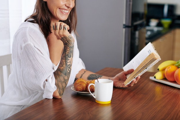 Giovane donna felice che legge un libro divertente quando mangia croissant e beve una tazza di caffè per colazione