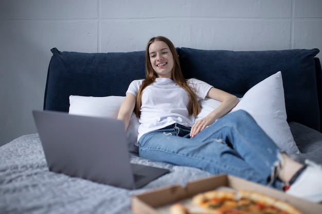 Giovane donna felice che mangia fetta di pizza calda a casa e guarda film sul computer portatile