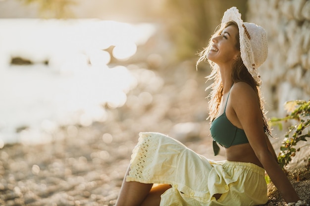 Giovane donna felice che si siede e che si diverte sulla spiaggia nel giorno d'estate.