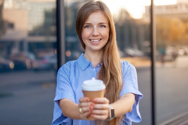 Giovane donna felice che sorride al caffè d'offerta della macchina fotografica in tazza di carta eliminabile