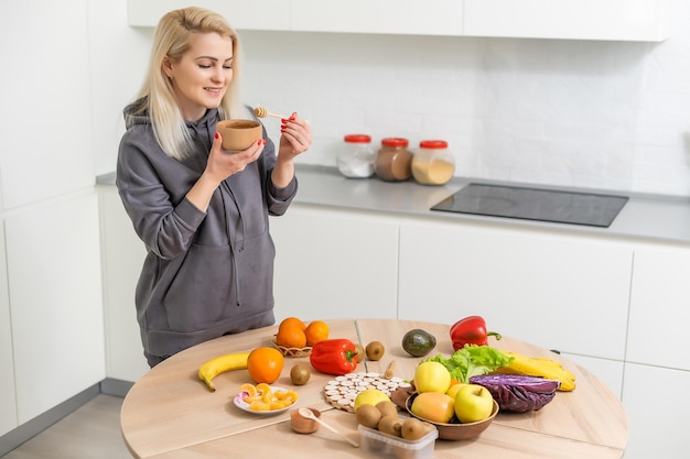 Giovane donna felice che tiene miele godendo durante la cottura di cibi sani in cucina. Dieta disintossicante, concetto di nutrizione dimagrante.