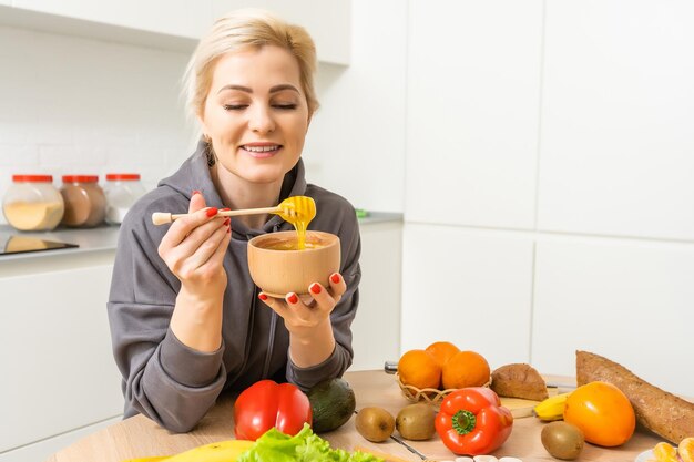 Giovane donna felice che tiene miele godendo durante la cottura di cibi sani in cucina. Dieta disintossicante, concetto di nutrizione dimagrante.