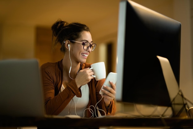 Giovane donna felice che usa il cellulare mentre beve il tè e lavora di notte a casa