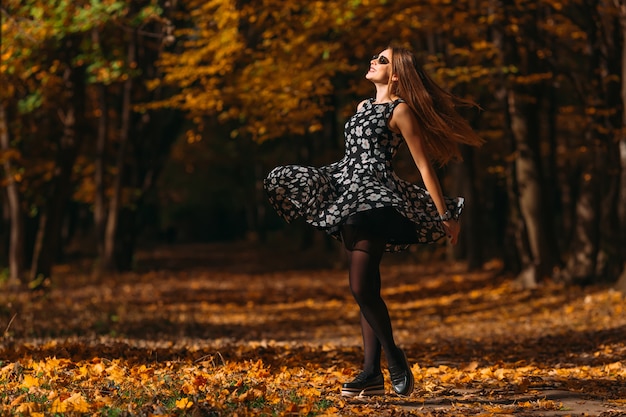 Giovane donna felice con gli occhiali da sole in un vestito nel parco di autunno che balla e che gira intorno con volare capelli lunghi e sorridere