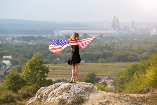 Giovane donna felice con i capelli lunghi alzando in su sventolando sulla bandiera nazionale americana vento nelle sue mani in piedi su un'alta collina rocciosa godendo una calda giornata estiva.