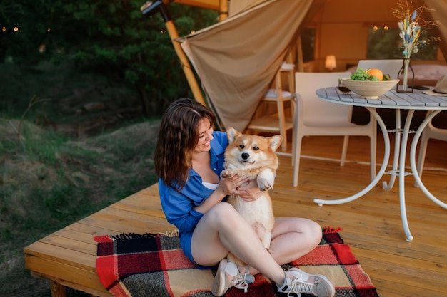 Giovane donna felice con il cane Welsh Corgi Pembroke che si rilassa nel glamping il giorno d'estate Tenda da campeggio di lusso per attività ricreative all'aperto e concetto di stile di vita ricreativo
