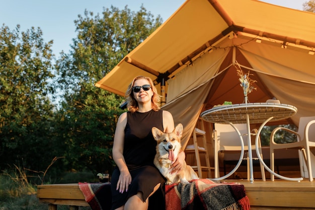 Giovane donna felice con il cane Welsh Corgi Pembroke che si rilassa nel glamping il giorno d'estate Tenda da campeggio di lusso per attività ricreative all'aperto e concetto di stile di vita ricreativo