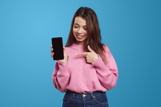 Giovane donna felice con il maglione rosa che sorride e guarda il cellulare mentre indica con il dito il cellulare