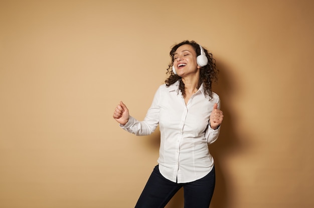 Giovane donna felice con le cuffie che ballano contro uno sfondo beige con spazio di copia