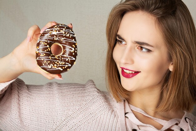 Giovane donna felice con le labbra rosa che tiene una ciambella al cioccolato su uno sfondo grigio