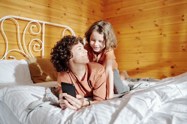 Giovane donna felice con lo smartphone e suo figlio che si rilassano sul letto