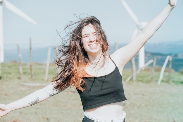 Giovane donna felice del ritratto frontale che gode della libertà alzando le braccia al cielo mentre sorride alla macchina fotografica circondata da mulini a vento elettrici che soffiano i capelli e si rilassano il concetto futuro della terra pulita