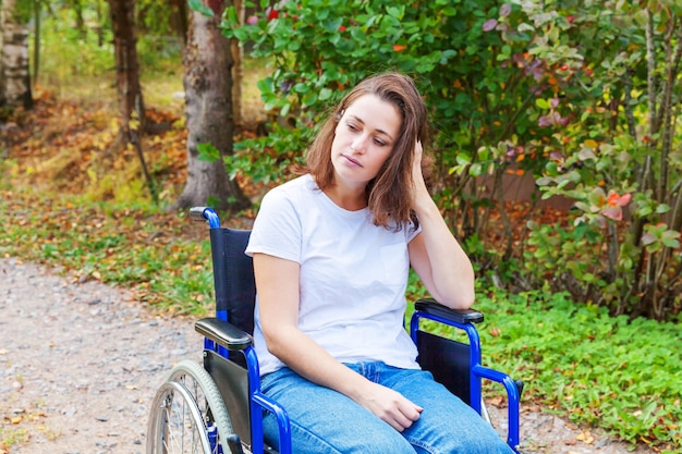 Giovane donna felice di handicap in sedia a rotelle sulla strada nel parco dell'ospedale che aspetta i servizi pazienti. Ragazza paralizzata in sedia invalida per disabili all'aperto in natura. Concetto di riabilitazione.