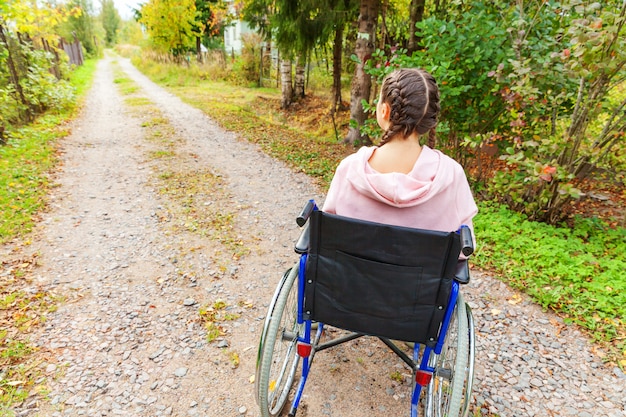 Giovane donna felice di handicap in sedia a rotelle sulla strada nel parco dell'ospedale che aspetta i servizi pazienti. Ragazza paralizzata in sedia invalida per disabili all'aperto in natura. Concetto di riabilitazione.