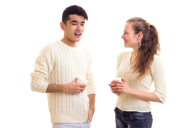 Giovane donna felice e giovane uomo bello in maglioni bianchi e jeans che tengono tazze bianche e parlano