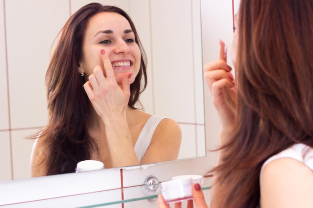 Giovane donna felice in camicia bianca con crema per il viso davanti allo specchio nel suo bagno