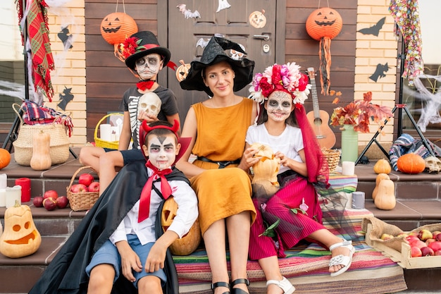 Giovane donna felice in cappello della strega e vestito giallo che si siede sulla scala dalla porta decorata della casa tra i bambini di halloween in costumi intelligenti