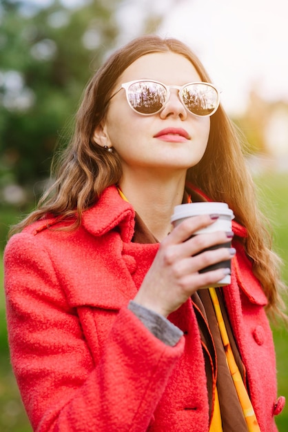 Giovane donna felice in cappotto rosso con caffè che cammina vicino agli alberi nel parco