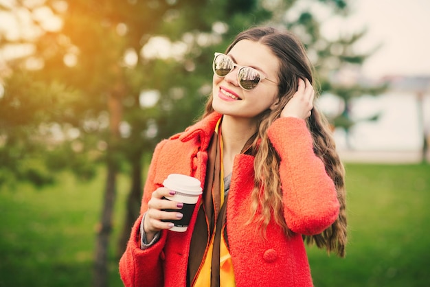 Giovane donna felice in cappotto rosso con caffè che cammina vicino agli alberi nel parco