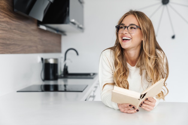 giovane donna felice in occhiali leggendo un libro e sorridendo alla cucina moderna