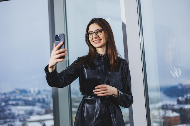 Giovane donna felice in una camicia nera che sorride e parla al telefono in un ufficio moderno con grandi finestre Lavoro a distanza