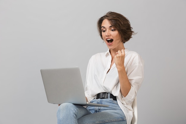 Giovane donna felice scioccata che si siede sullo sgabello isolato sopra il computer portatile della tenuta della parete grigia