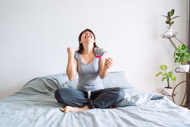 Giovane donna felice seduta sul letto che guarda il test di gravidanza positivo