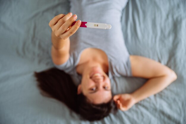 Giovane donna felice seduta sul letto che guarda la vista dall'alto del test di gravidanza positivo