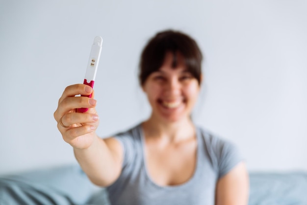 Giovane donna felice seduta sul letto guardando il test di gravidanza positivo sfondo sfocato