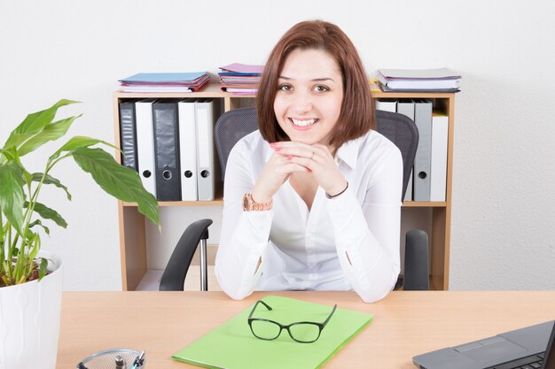 Giovane donna felice sorridente che si siede al suo scrittorio in ufficio