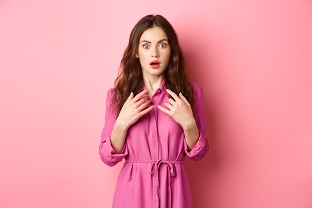 Giovane donna femminile scioccata ansimante, tenendo le mani sul petto e fissando senza parole la telecamera, guarda con incredulità, in piedi contro il muro rosa.