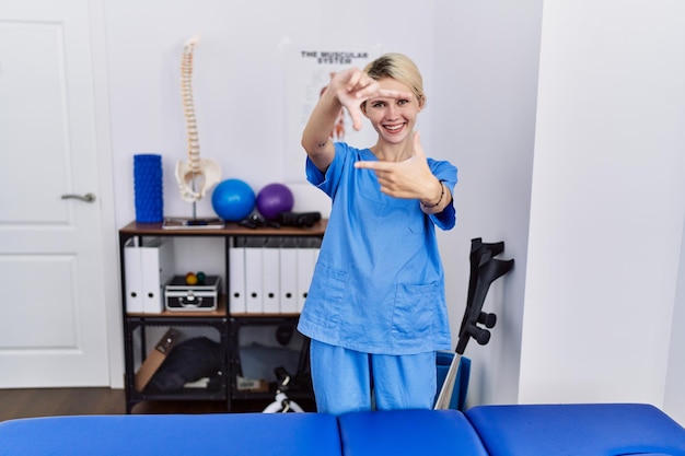 Giovane donna fisioterapista che lavora presso la clinica per il recupero del dolore che sorride facendo cornice con le mani e le dita con la faccia felice. concetto di creatività e fotografia.