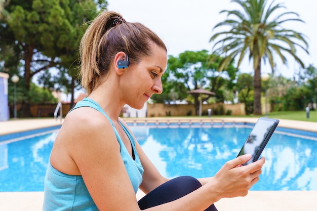 Giovane donna fitness che sceglie una lezione di sport online all'aperto. Vista laterale orizzontale della giovane donna che si connette con la tecnologia a una lezione di ginnastica con gli amici accanto a una piscina.