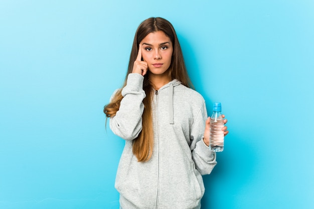 Giovane donna fitness in possesso di una bottiglia d'acqua che punta il suo tempio con un dito, pensando, incentrato su un compito.