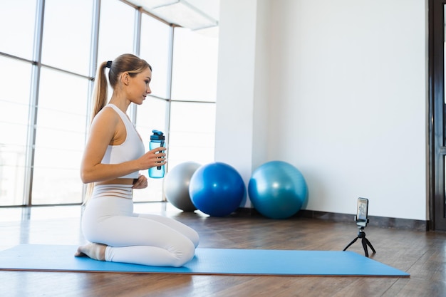 Giovane donna fitness seduta sul tappetino yoga con bottiglia d'acqua e allenamento in streaming dal vivo