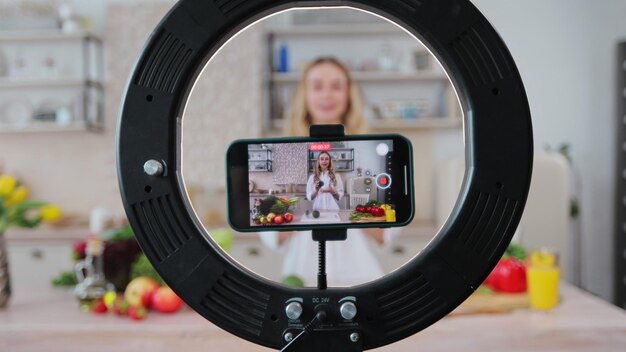 Giovane donna food blogger che cucina insalata davanti alla fotocamera dello smartphone mentre registra video vlog e live streaming a casa in cucina