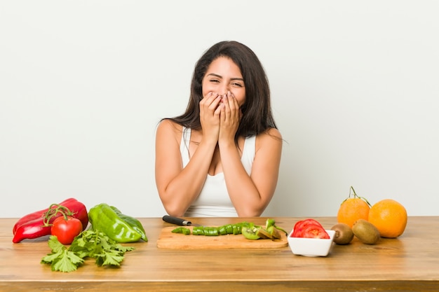 Giovane donna formosa che prepara un pasto sano ridendo di qualcosa, coprendo la bocca con le mani.