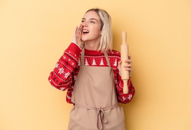 Giovane donna fornaia caucasica che fa i biscotti per Natale isolata su sfondo giallo gridando e tenendo il palmo vicino alla bocca aperta.