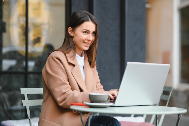 Giovane donna freelance che si gode un caffè mentre lavora all'aperto su un computer portatile collegato al Wi-Fi pubblico. Elegante studentessa che studia online in città.