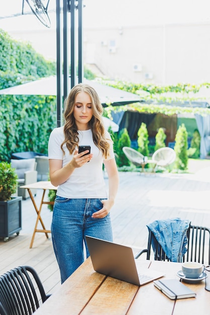 Giovane donna freelance in abiti casual che beve caffè e parla al telefono lavorando su una terrazza verde guardando uno schermo e lavorando a un progetto a distanza dal lavoro in un bar