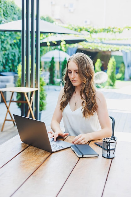 Giovane donna freelance positiva seduta a un tavolo con un laptop e che lavora su una terrazza verde guardando lo schermo e lavorando a un progetto da remoto in un caffè